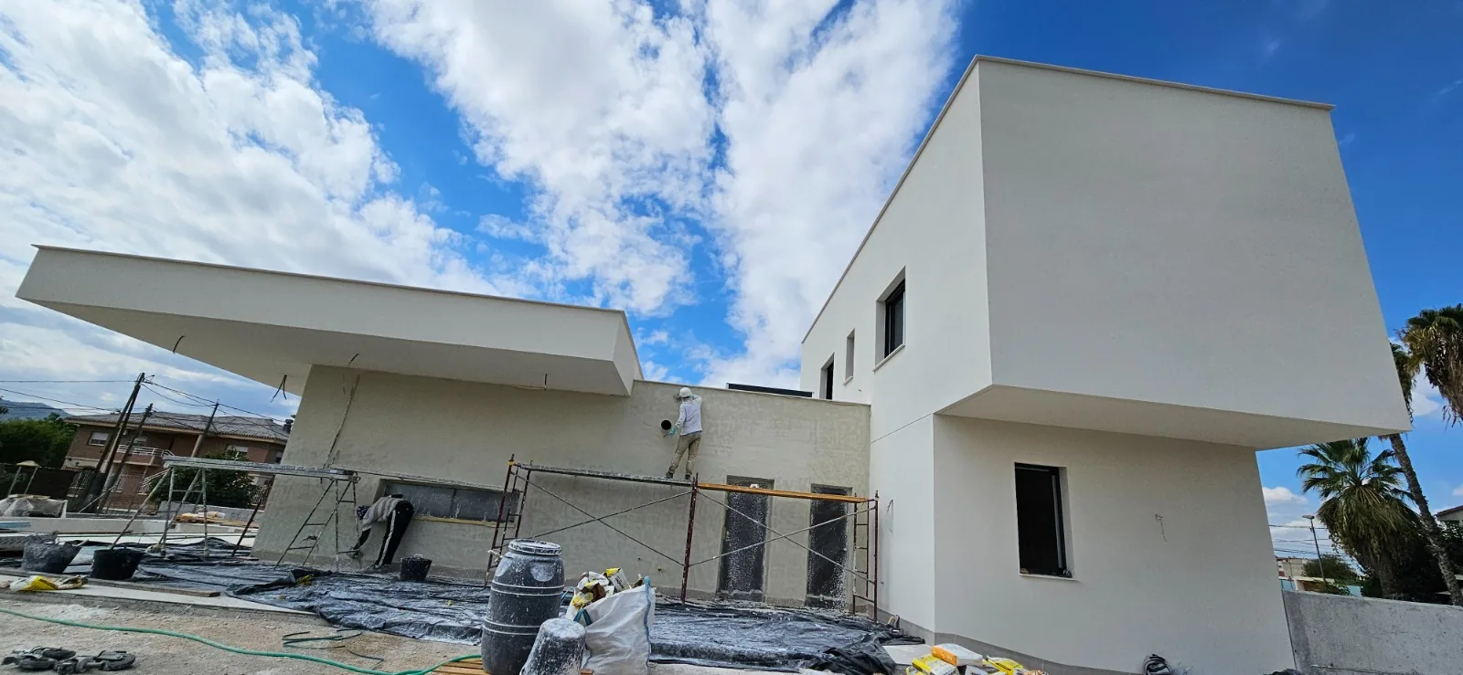 Operario aplicando revestimiento en fachada blanca de vivienda de dos alturas con cielo despejado